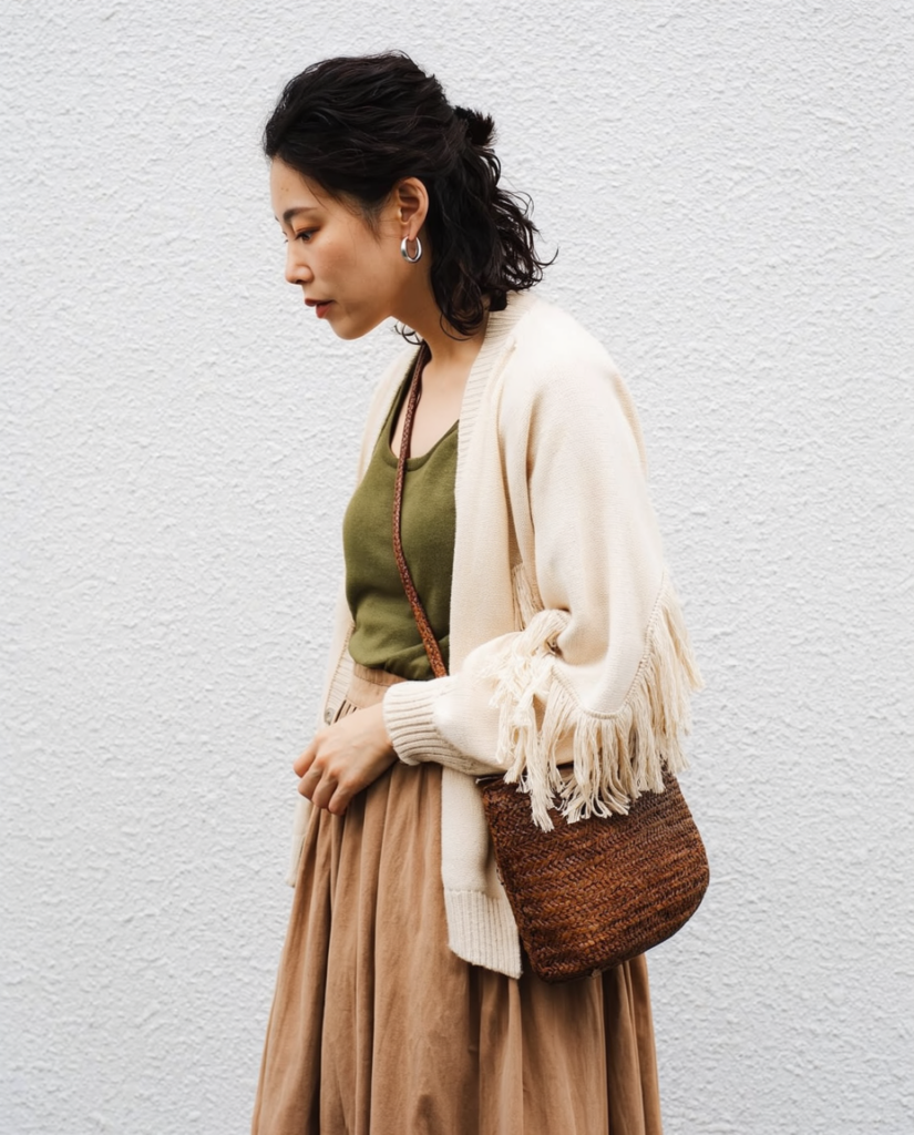 Woman in an olive green top with a beige fringed cardigan and tan skirt carrying a brown woven crossbody bag