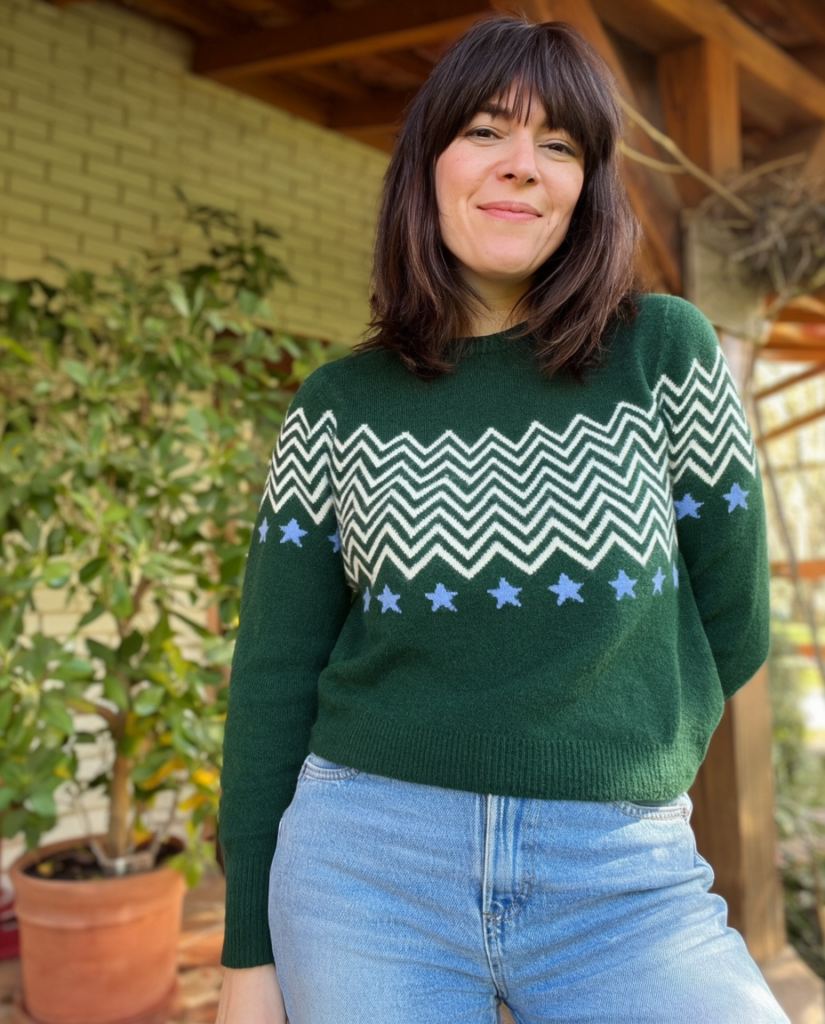 Woman in a green patterned sweater smiling outdoors standing beside potted plants in a garden setting
